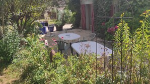 A garden area with a round table and chairs, surrounded by various plants and a stone building with red shutters. - Lac Aigueblette Maison de Charme (Nances)