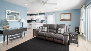 A living room with a brown leather sofa, a ceiling fan, a small table, and a kitchen in the background. - Awesome condo with pool, across from beach access (Navarre)