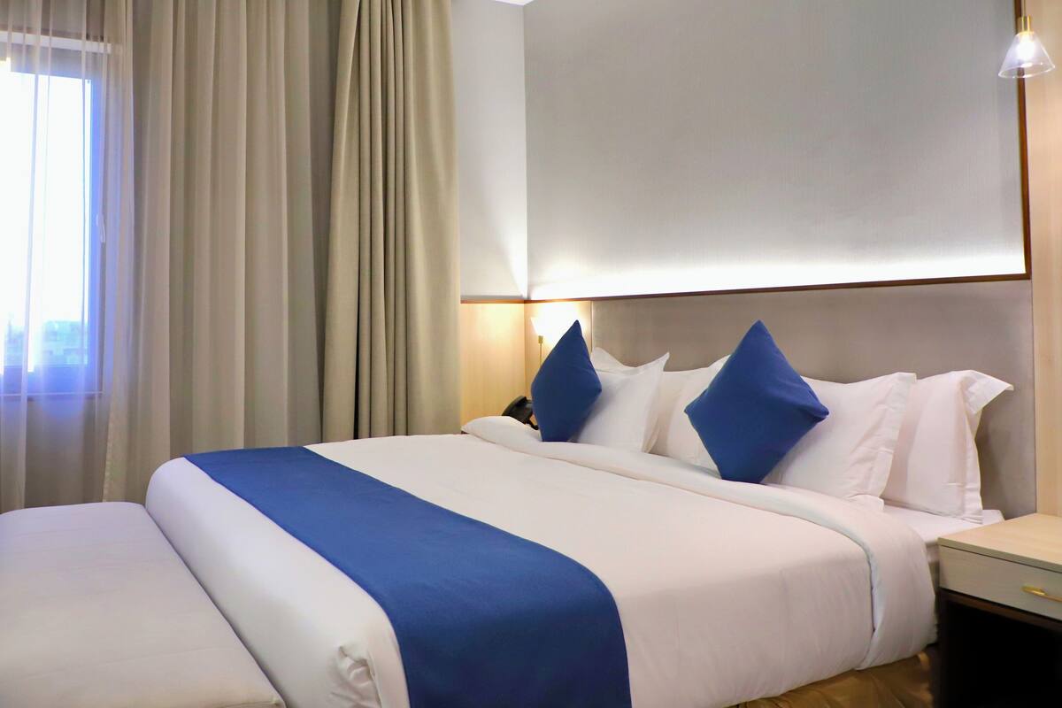 A hotel room with a large bed, a bedside table, a lamp, and a view of the city through the window.