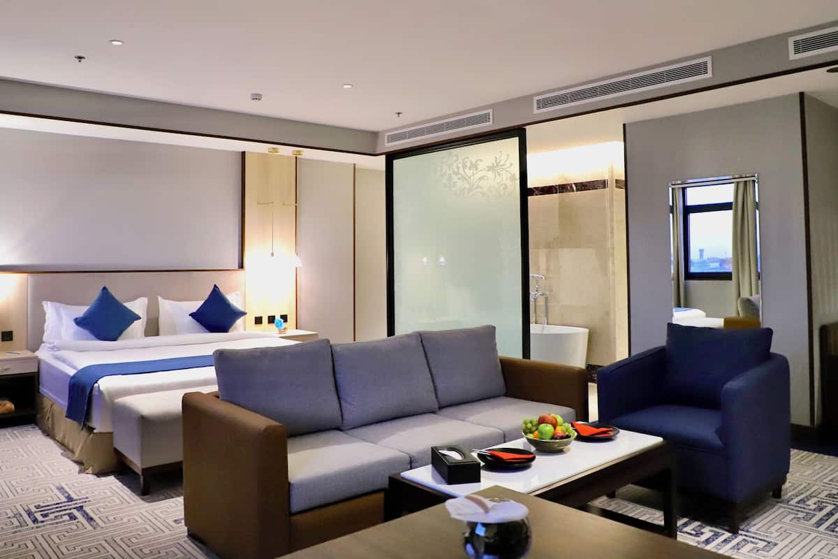 A modern hotel room with a bed, a grey sofa, a blue armchair, a small table with a bowl of fruit, and a large mirror.