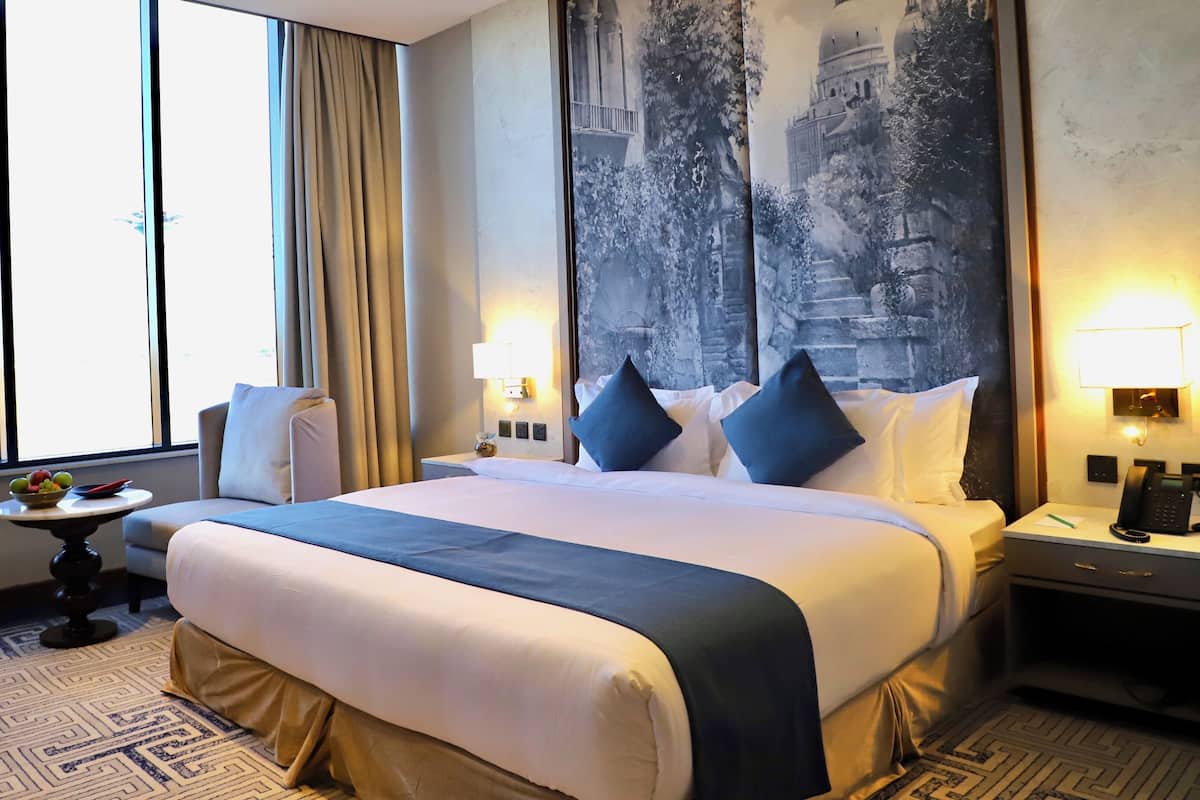 A hotel room with a large bed, a small table with fruit, a nightstand with a phone, and a wall with a historical mural.