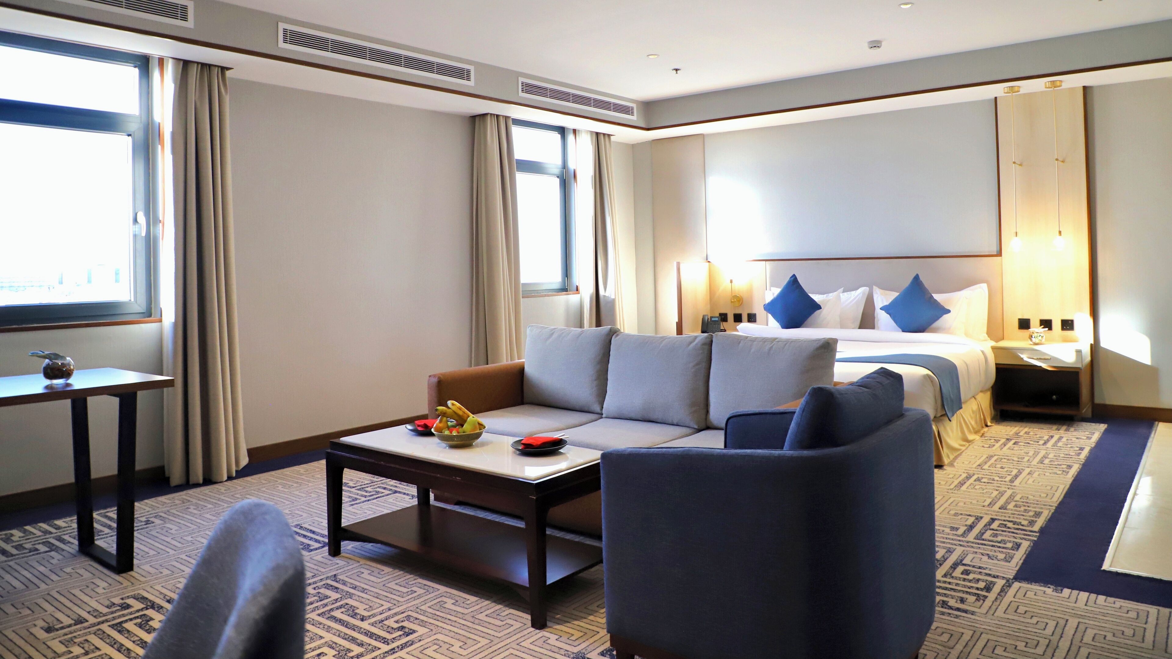 Habitación de hotel con sofá, mesa de centro, cama con almohadas azules, mesita de noche con lámpara y ventana con cortinas.