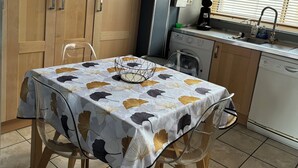 Una cocina con armarios de madera, una mesa de comedor con mantel estampado de hojas, una lámpara colgante y un ventanal con persianas.