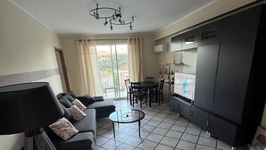 Una sala moderna con sofá, una mesa de centro y un comedor con sillas. Se observa un amplio ventanal con cortinas, y en el fondo, una zona de cocina.