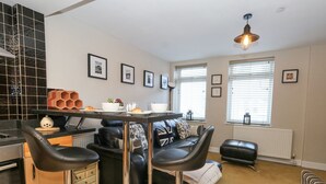 A modern kitchen with a dining area, featuring a black leather sofa, two bar stools, and a table set for two. - Anchor Cottage (Weymouth)