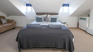 A bedroom with a large bed, two windows, a radiator, a dresser, and a brown leather sofa. - Anchor Cottage (Weymouth)