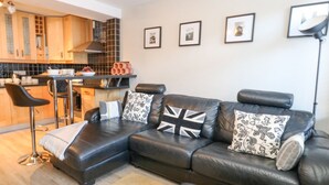 A modern living room with a black leather sofa, a small kitchen area, and a dining bar. - Anchor Cottage (Weymouth)