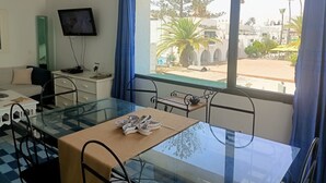 A room with a glass table, chairs, a television, and a view of a courtyard with palm trees and buildings. - Magnificent S+1 with panoramic view of Port El Kantaoui (Hammam Sousse)