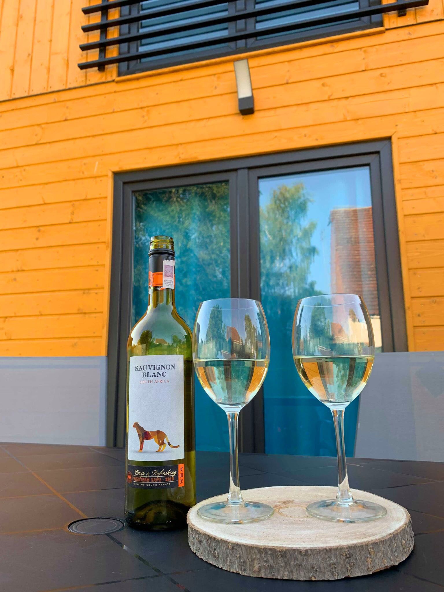 A bottle of Sauvignon Blanc and two glasses on a round stone table outside a building with yellow siding.