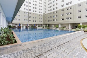 Une piscine entourée d’une terrasse dallée de pierres, avec un immeuble à plusieurs étages en arrière-plan.