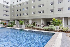 A swimming pool with tiled flooring, surrounded by a low stone wall and various plants. In the background, there is a multi-story building with balconies and windows. - Capital O 93593 Pelangi Loftville City (South Tangerang)