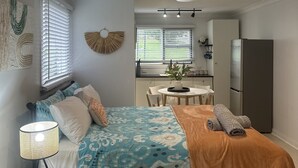 Un espace de vie compact comprenant un lit, une petite table, une lampe, un réfrigérateur et un coin cuisine à l’arrière-plan.