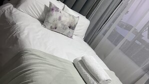 Un lit bien fait, avec une literie blanche et grise, un coussin à motifs floraux et une serviette pliée.