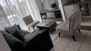 Un salon moderne avec un canapé, un fauteuil et une table à manger. Un téléviseur à écran plat est fixé au mur, et on aperçoit un coin cuisine au fond de la pièce.