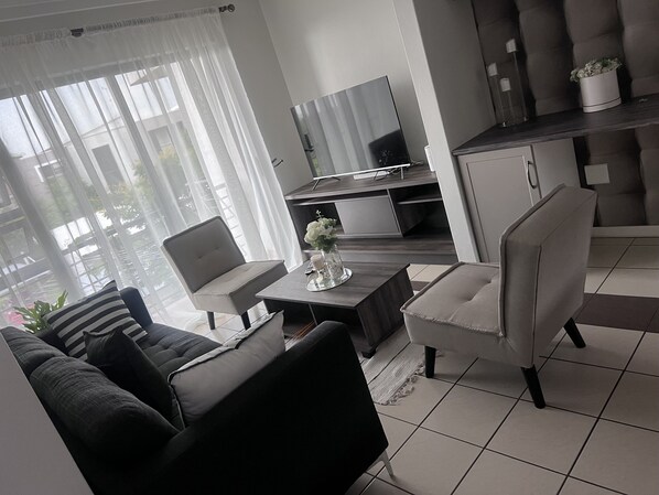 A modern living room with a sofa, armchair, and a dining table. A flat-screen TV is mounted on the wall, and there is a kitchen area in the background.