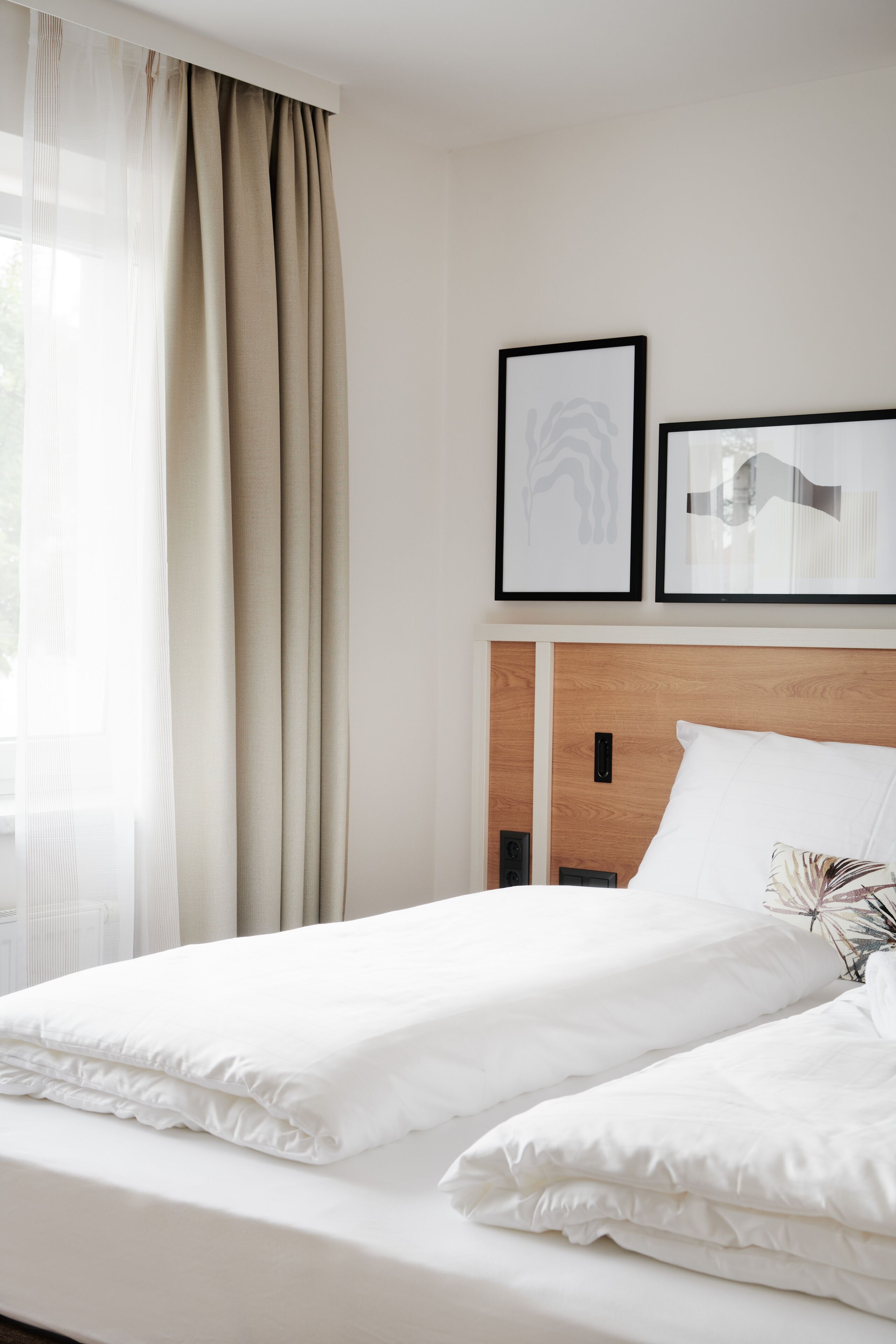 A neatly made bed with white linens, a wooden headboard, and two framed artworks above it.
