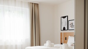 A modern hotel room with a large bed, wooden flooring, and a view of the outdoors through sheer curtains.