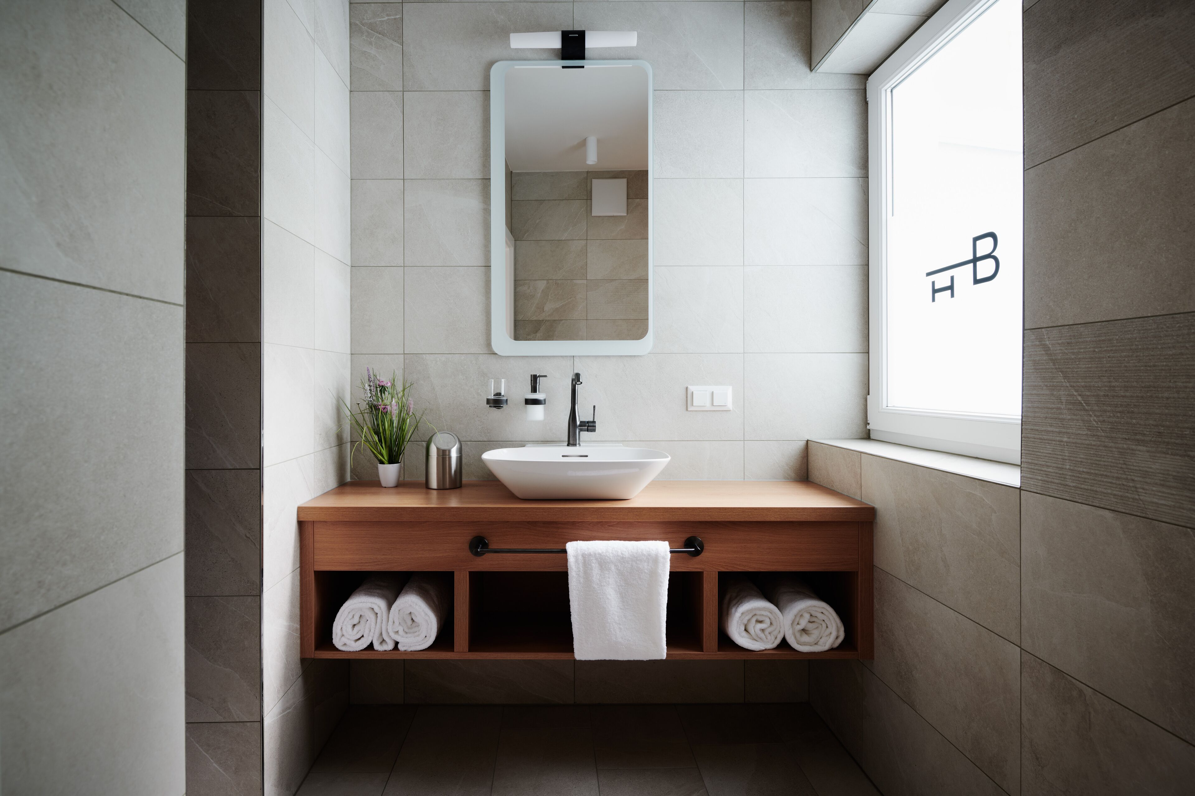 Une salle de bain moderne dotée d’un grand miroir, d’un lavabo sur colonne, d’un meuble-lavabo en bois, d’un espace de rangement pour les serviettes et d’une fenêtre offrant une belle vue.