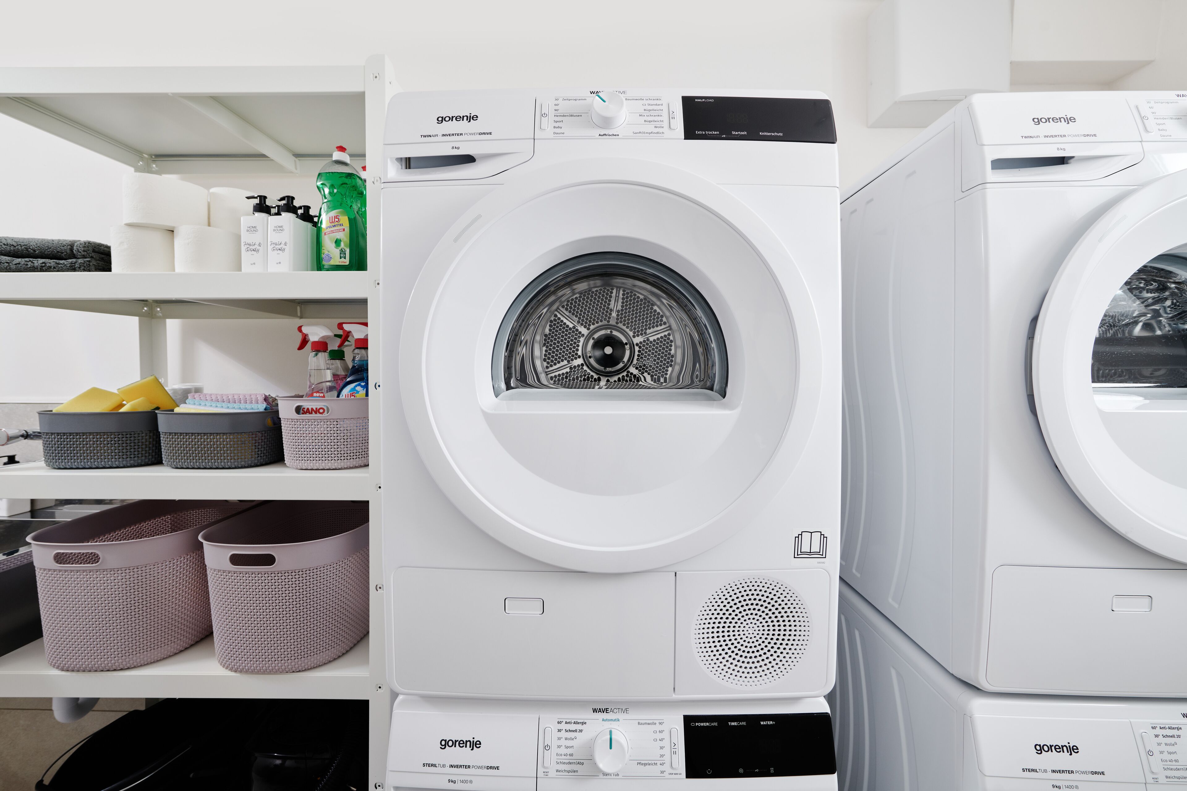 A row of white washing machines with control panels and a shelf above containing laundry supplies.