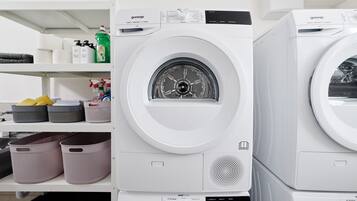 A row of white washing machines with control panels and a shelf above containing laundry supplies.