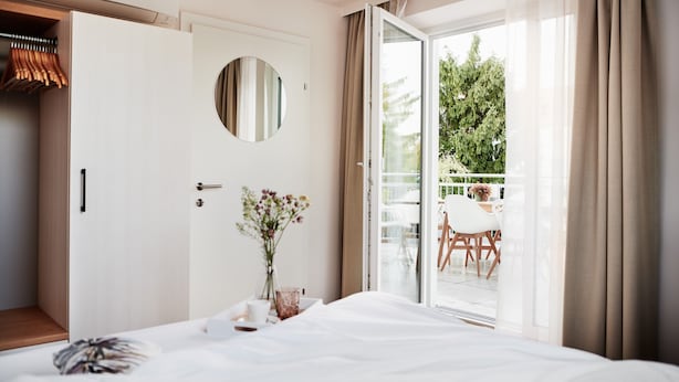 A hotel room with a bed, a round mirror, a vase with flowers, a wooden wardrobe, and a sliding glass door to a balcony with outdoor furniture.