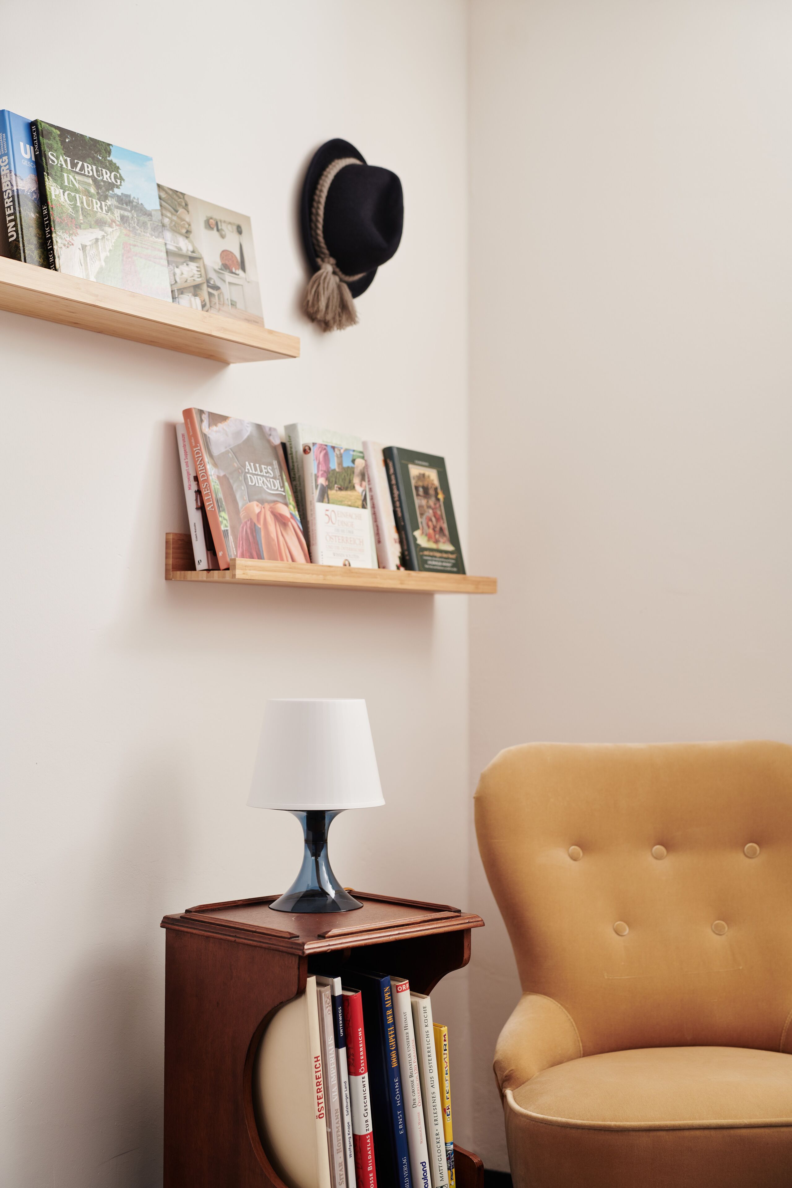 Una habitación con una mesita de madera que sostiene libros, una lámpara y un sombrero colgado en la pared.