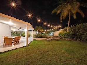 Un patio moderno al aire libre con comedor, luces colgantes y abundante vegetación.