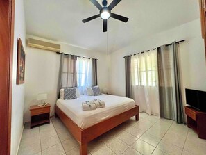 Un dormitorio con cama, mesita de noche, mueble para televisor, ventilador de techo y cortinas.