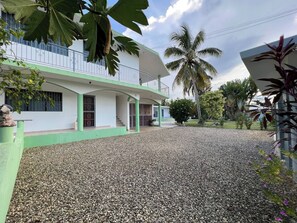 Un bâtiment à deux étages doté d’un balcon, d’une allée en gravier et d’une végétation tropicale.