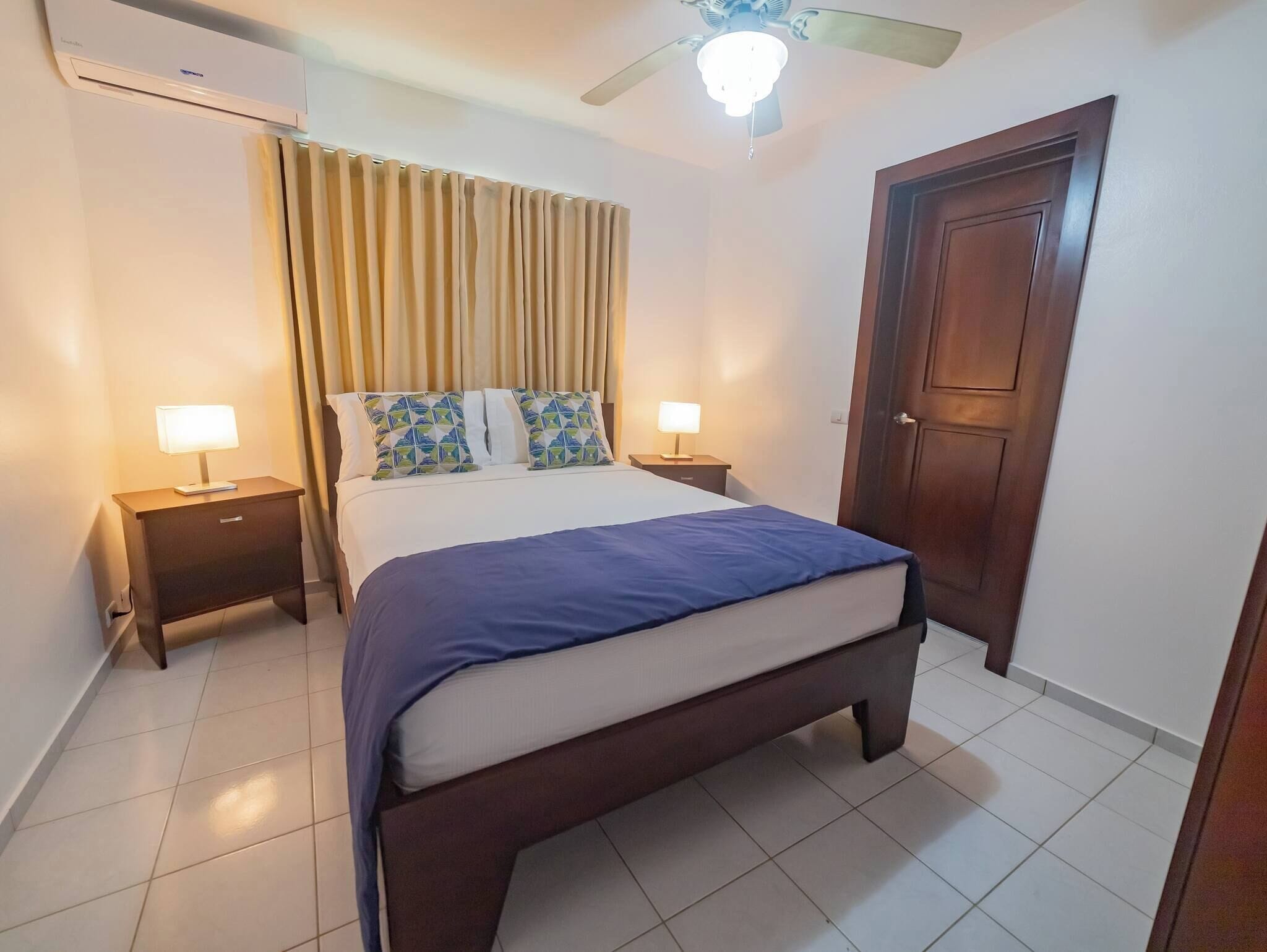 A bedroom with a bed, two bedside tables with lamps, a ceiling fan, a door, and an air conditioning unit.