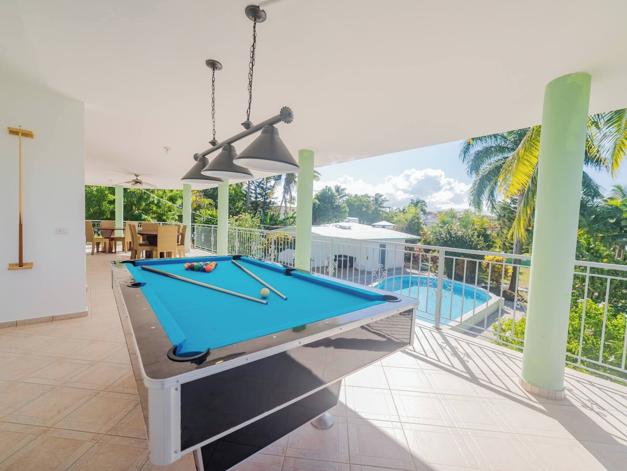A pool table with a blue felt surface, a ceiling fan, and a view of a swimming pool and greenery.