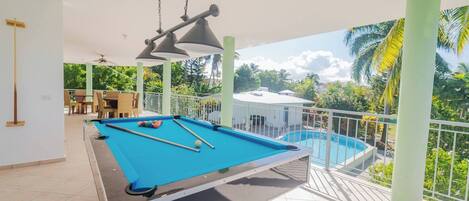 A pool table with a blue felt surface, a ceiling fan, and a view of a swimming pool and greenery.