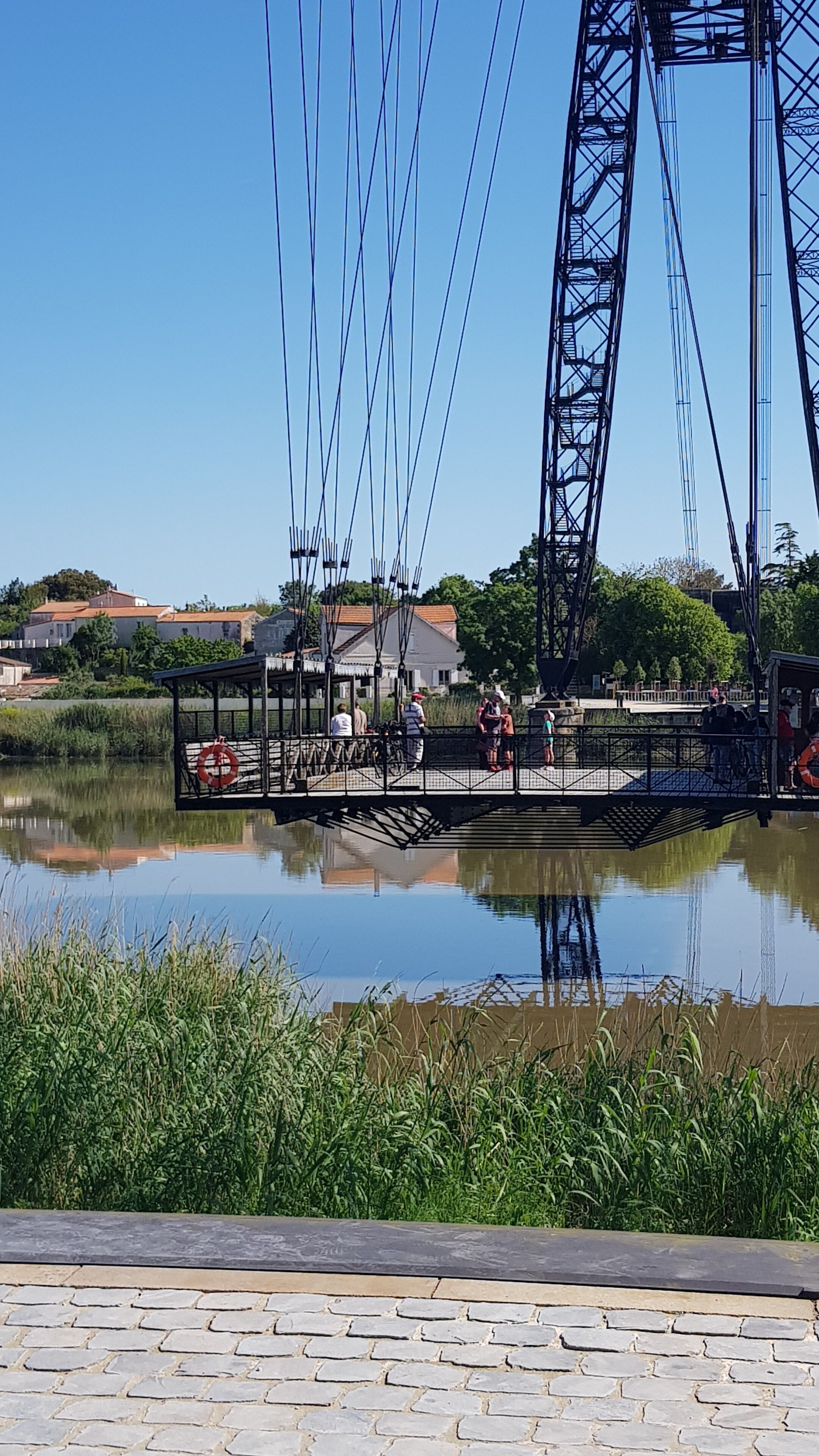 A bridge with a pedestrian walkway over a body of water, surrounded by greenery and buildings in the background.