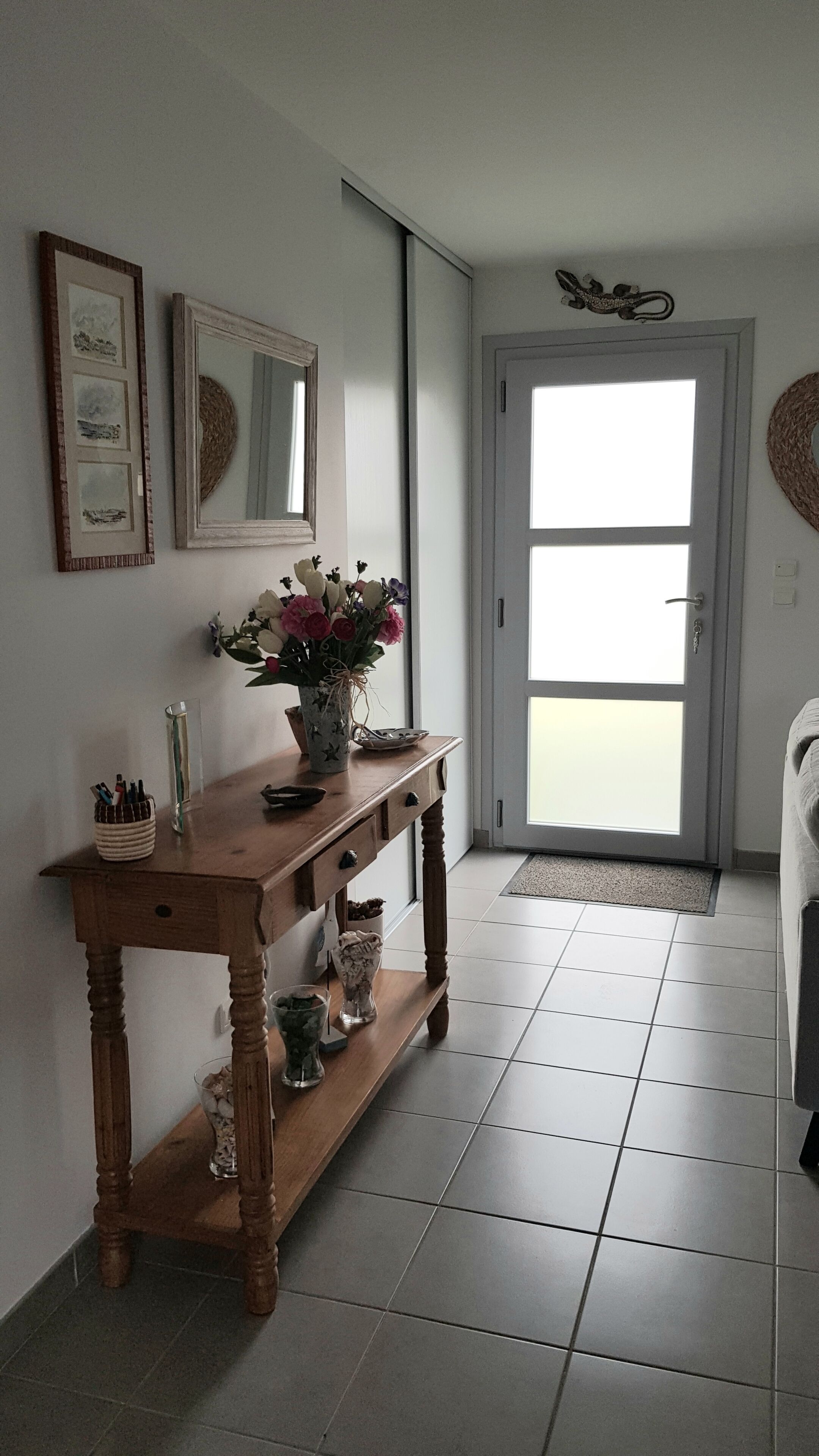 A hallway with a wooden console table, a vase with flowers, framed pictures, a mirror, and a door leading to a bright area.