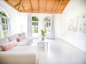 Un salon moderne avec un plafond en bois, un canapé avec des coussins, une table basse ornée d’un vase et de grandes fenêtres avec des rideaux.