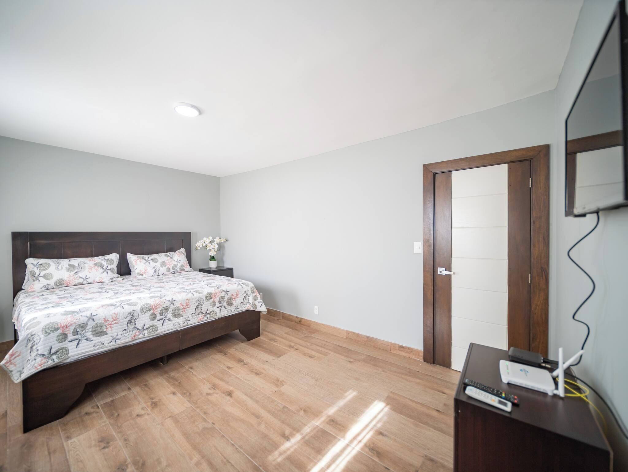 A bedroom with a bed, wooden flooring, a nightstand with a vase of flowers, a dresser with a mirror, and a door.