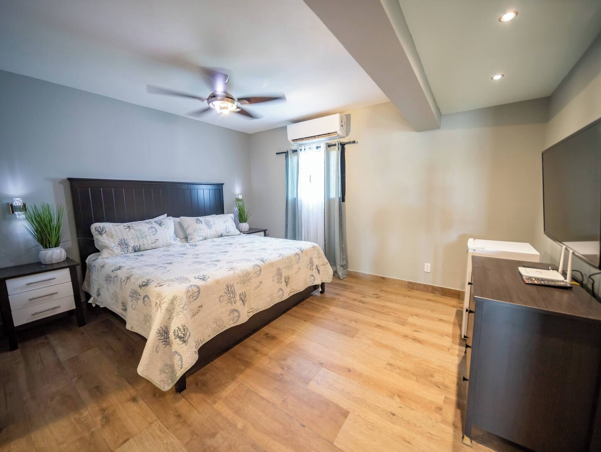 Ein Schlafzimmer mit Bett, Nachttischen, Schreibtisch, Deckenventilator, Klimaanlage und Fernseher.