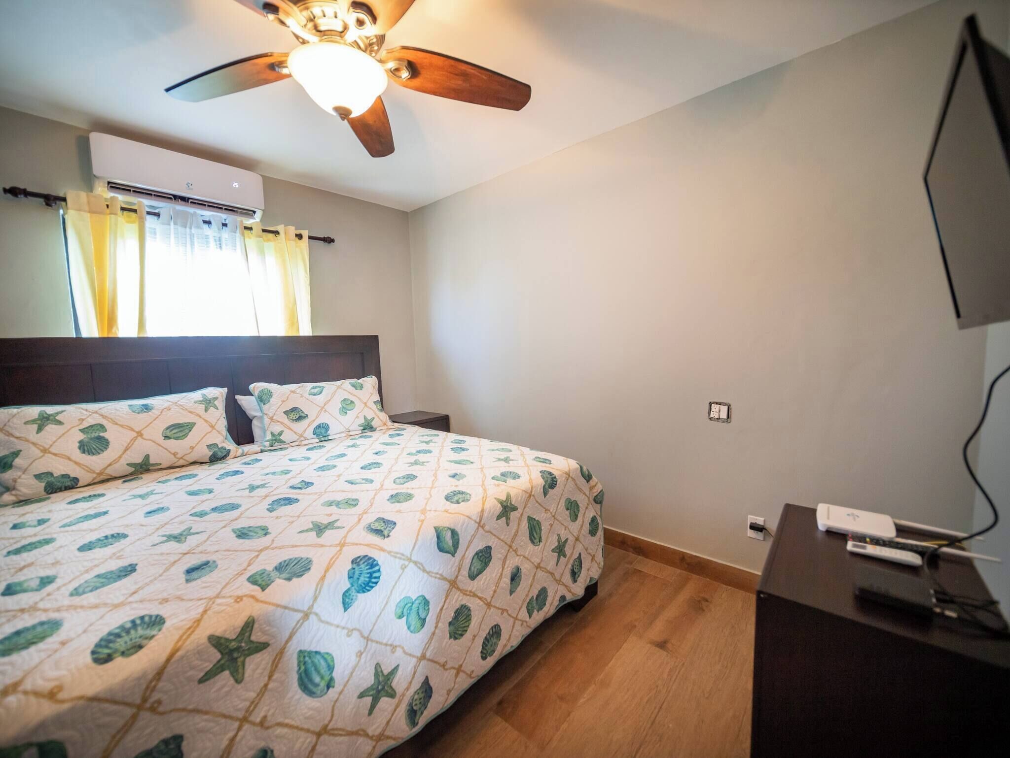 Een slaapkamer met een bed, een plafondventilator, een airconditioningapparaat, een nachtkastje, een bureau met een computer en een tv aan de muur.