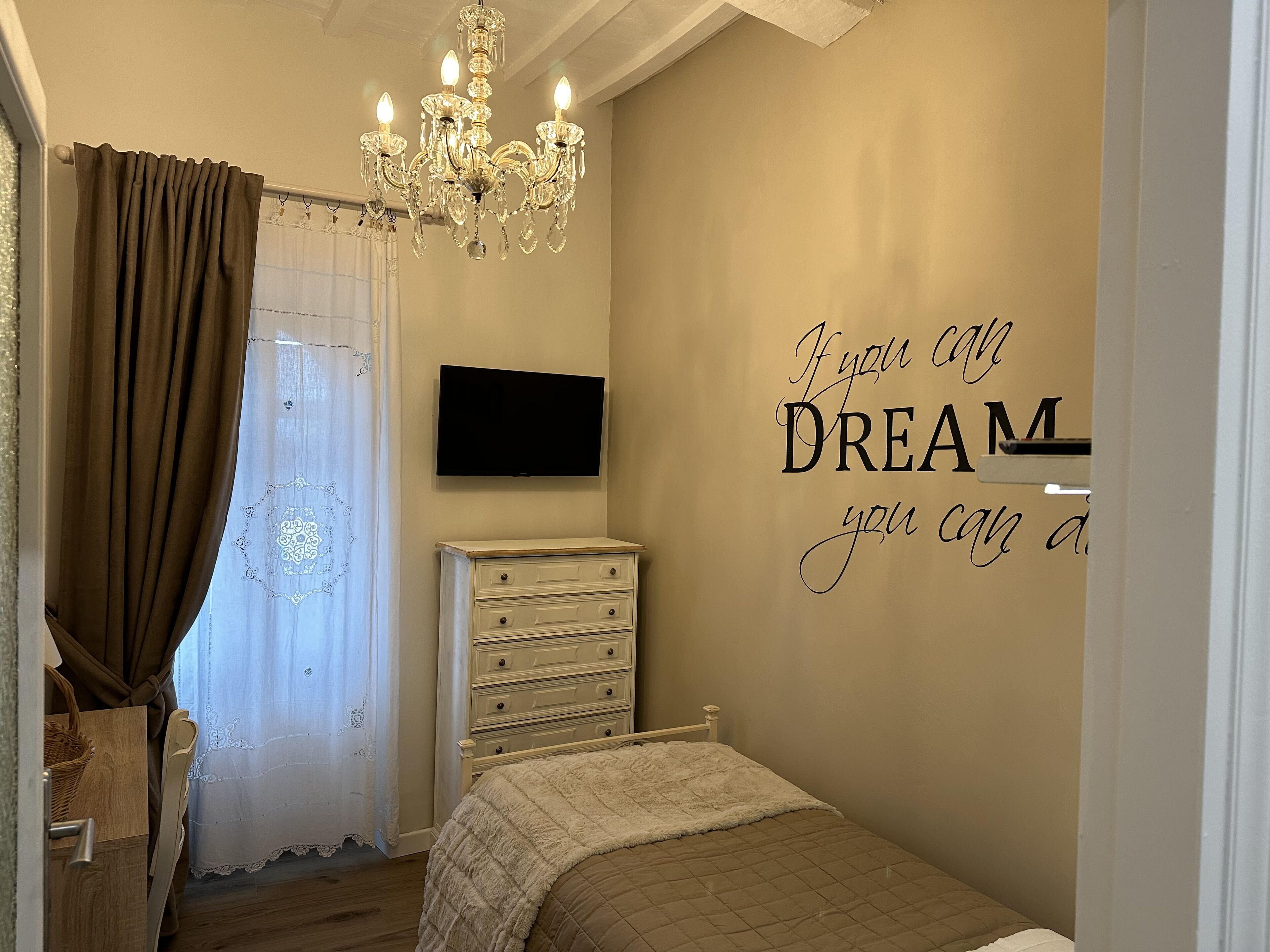 Un dormitorio con cama, cómoda, televisor, araña y una frase motivacional en la pared.
