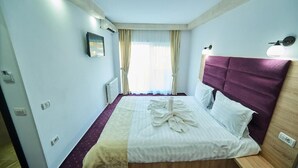 Habitación de hotel con una cama, cabecera, mesita de noche, ventana con cortinas y una lámpara fijada a la pared.