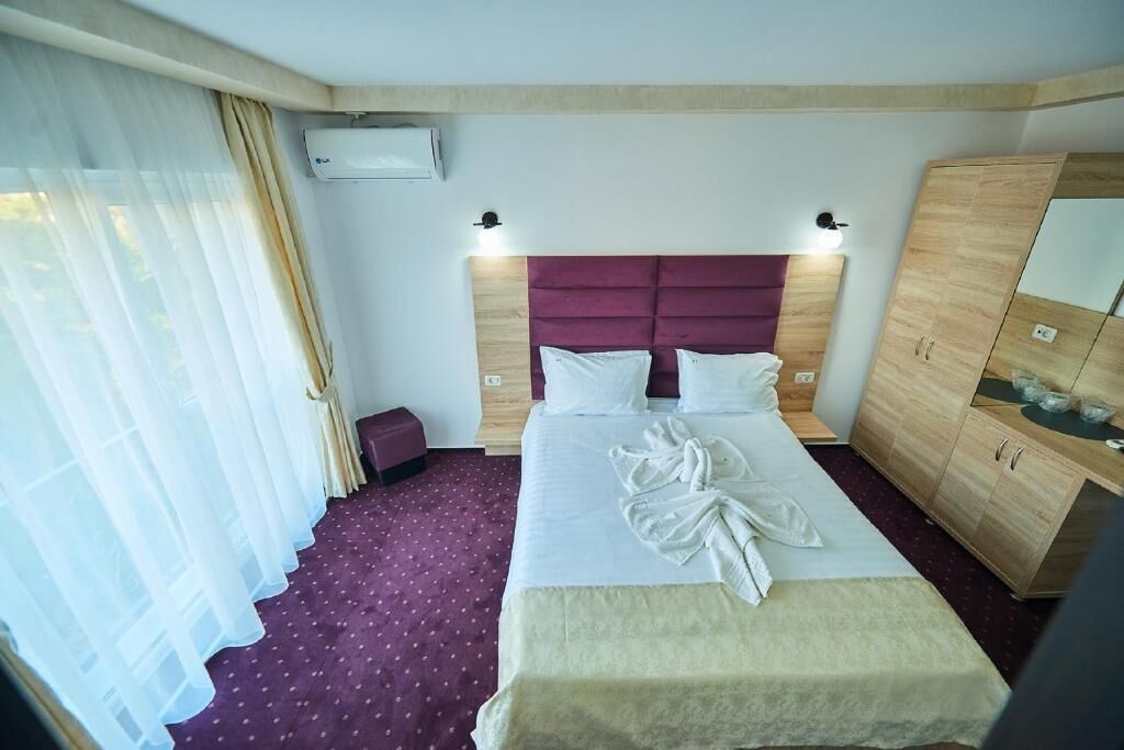 A hotel room with a large bed, a wooden wardrobe, a small bedside table, and a window with sheer curtains.