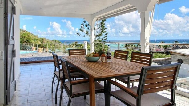 Un espace extérieur abrité, aménagé pour prendre les repas, avec une table et des chaises en bois, un saladier rempli de fruits et une bouteille de spiritueux.