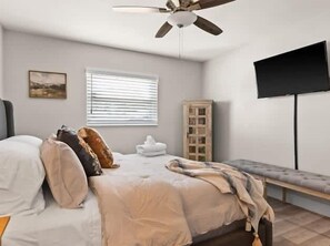 A bedroom with a bed, a ceiling fan, a window with blinds, a television mounted on the wall, and a wooden cabinet.