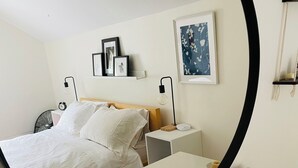 Ein ordentlich eingerichtetes Hotelzimmer mit Bett, Nachttischen, einem Spiegel und einem an der Wand befestigten Regal mit gerahmten Bildern.