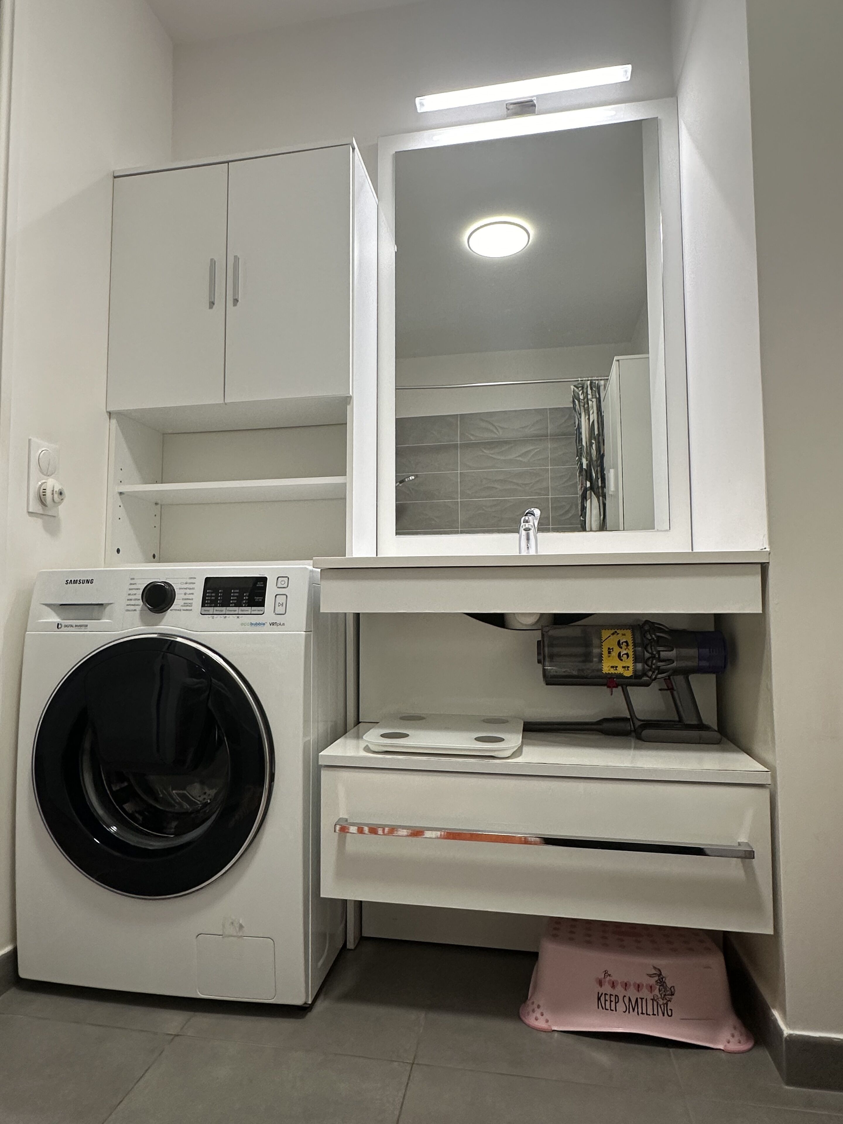 A compact laundry area with a washing machine, a mirror above the sink, and a small countertop with a bottle and a cloth.