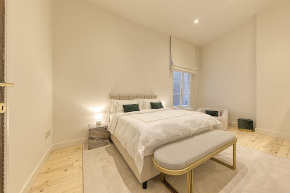 A bedroom with a large bed, bedside tables, a bench, a window, and a small chair.