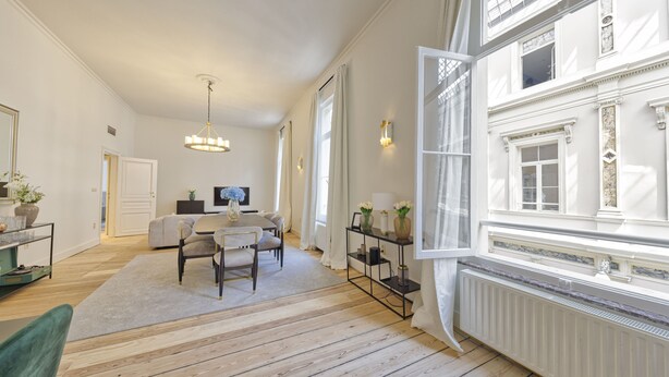 Ein geräumiges Wohnzimmer mit Holzboden, einem Essbereich mit Tisch und Stühlen, einem großen Fenster mit Vorhängen und einem Heizkörper.