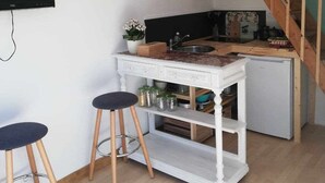 Eine kleine Küche mit weißer Arbeitsplatte, zwei Barhocker, einem kleinen Kühlschrank und einer Holztreppe.