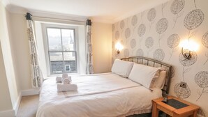 A neatly made bed with white linens, a bedside table with a lamp, and a window with curtains. - La'l Tethera (Ambleside)