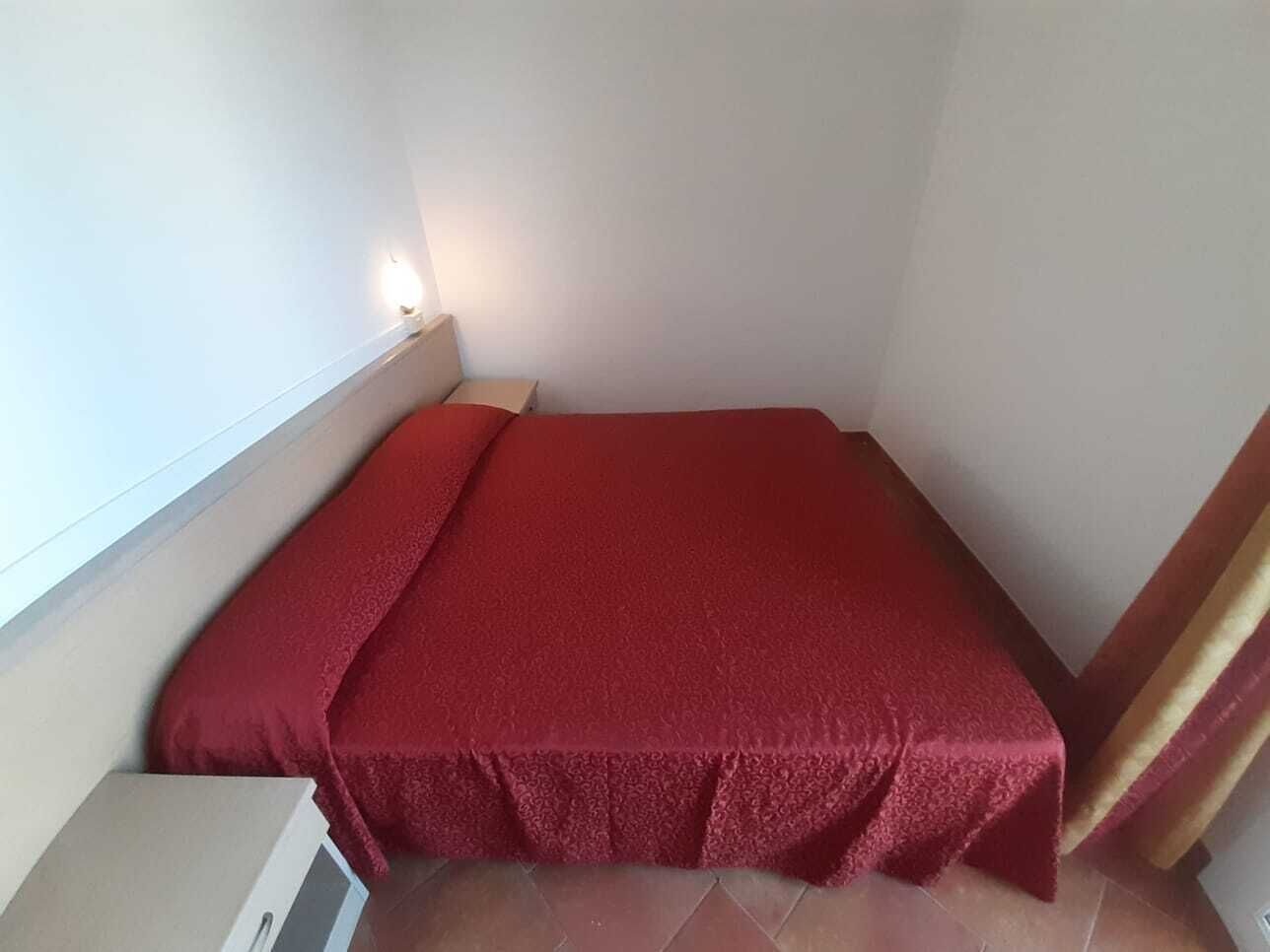 Una cama pequeña y ordenada con una colcha roja en una habitación de esquina con paredes blancas y suelo de baldosas.